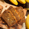 Loaf of banana bread with slices on a wooden cutting board, surrounded by bananas and pecans.