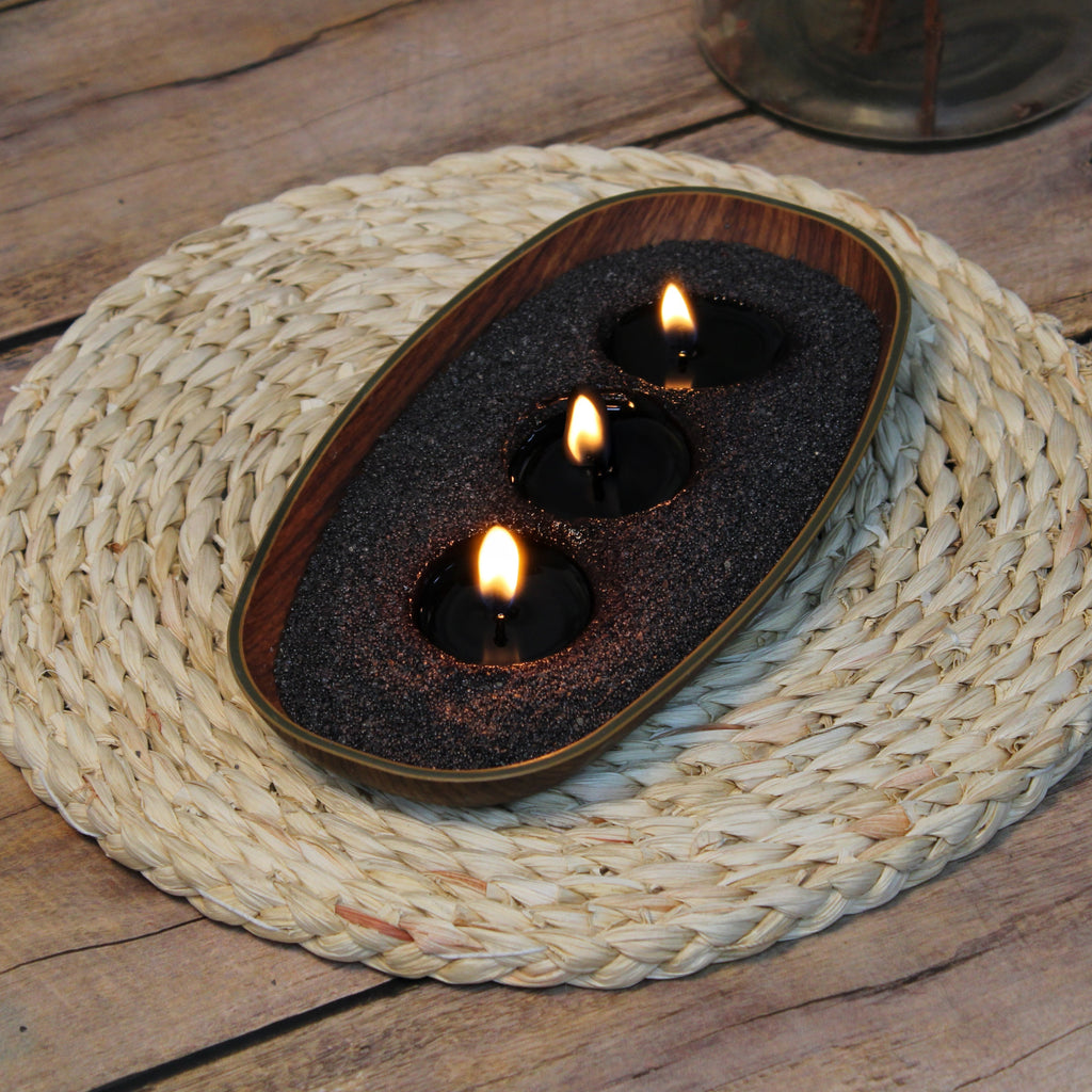 Dark pearled candle wax in a wooden bowl with lit wicks on a woven mat